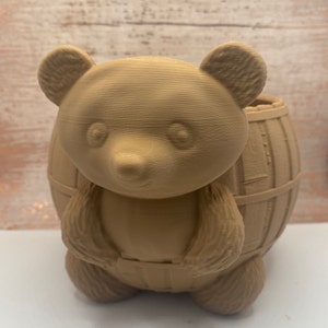 May include: A brown ceramic planter shaped like a bear wearing overalls. The bear is holding a barrel on its back.