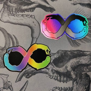 Op de afbeelding: Twee stickers in de vorm van een oneindigheidssymbool met een zwarte achtergrond en een regenboogkleurige gradiënt. De stickers hebben een druppelend verf-effect en een spookachtig figuur aan elke kant.