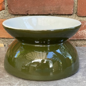 May include: A two-tiered ceramic bowl with a glossy olive green exterior and a white interior. The bowl has a rounded base and a wider, shallow top. The surface reflects the surrounding environment.