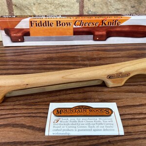 May include: A wooden fiddle bow cheese knife with a metal blade. The knife is made by Mountain Woods and comes with a small card that says "Thank you for purchasing Mountain Woods' Fiddle Bow Cheese Knife. You will find this knife ideal for use with our Fiddle Cutting Board or Cutting Center. Each of our finely crafted products is guaranteed against defective workmanship."