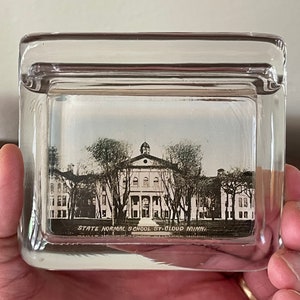 May include: A clear, rectangular glass paperweight with a black and white image of the State Normal School St. Cloud Minn. The image is of a large building with columns and trees. The paperweight is held in a hand.
