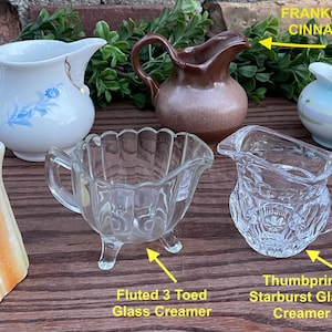 May include: A collection of small pitchers, including a brown and white pitcher labeled "Frankoma Brown or Cinnamon Pitcher 5 inches", a white pitcher with blue floral designs, a blue and white pitcher with floral designs, a clear glass pitcher with a thumbprint design, a clear glass pitcher with a fluted design and three feet, and a brown and white pitcher with a wood-grain design.
