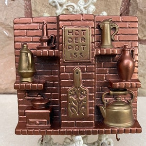May include: A decorative wall hanging featuring a brick-like design with miniature kitchen items. The piece includes a kettle, pitcher, and a sign that reads "HOT DEPOT ISS". The color scheme is primarily bronze and brown.