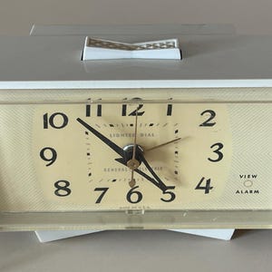 May include: A vintage General Electric alarm clock with a cream-colored face and black numerals. The clock has a white plastic casing and a clear plastic cover. The words "LIGHTED DIAL" and "VIEW ALARM" are visible on the face.