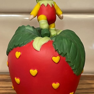 May include: A red ceramic strawberry with yellow hearts and green leaves. A small ceramic elf figure stands on top of the strawberry.