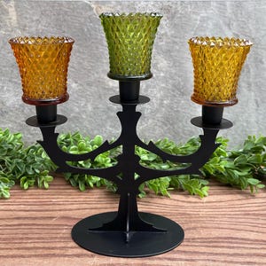 May include: A black metal candle holder with three arms, each holding a glass candle holder. The candle holders are decorated with a diamond pattern and are in shades of green and amber.