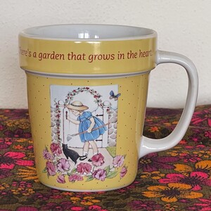 May include: Yellow ceramic mug with a white handle. The mug features a garden scene with a girl, a black cat, and flowers. The text "There's a garden that grows in the heart" is printed on the rim. The mug has a dotted pattern.