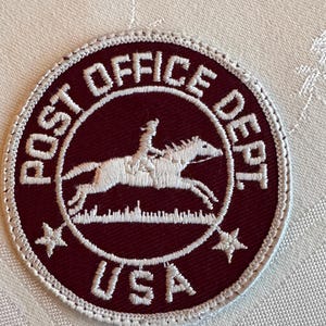 May include: A circular embroidered patch featuring a deep maroon background with white stitching. The center depicts a rider on a galloping horse above a grassy line. The text reads POST OFFICE DEPT and USA, with two five-pointed stars at the bottom.