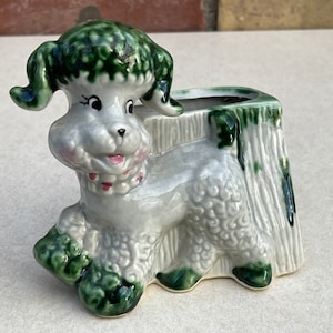 May include: A ceramic poodle planter in a whimsical design. The poodle is light gray with green accents on its ears, paws, and the top of the planter. It has a pink mouth and a red collar. The planter is shaped like a tree stump.
