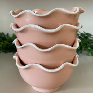 May include: A stack of four pink ceramic bowls with a ruffled edge and white trim. The bowls are stacked on top of each other, with the top bowl slightly angled. The bowls have a smooth, glossy finish and are likely for serving food.