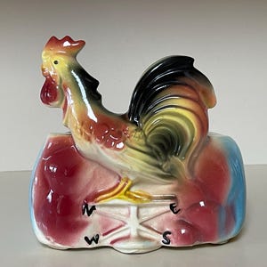 May include: A ceramic rooster figurine with a colorful design. The rooster has a red comb and wattle, with yellow, black, and orange feathers. The base is painted with red, blue, and white, and features a compass rose with the letters N, E, S, and W.