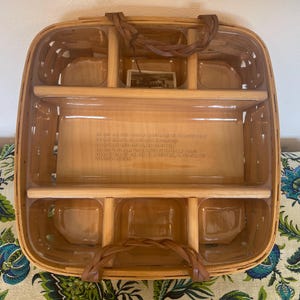 May include: A square woven wood basket featuring a clear plastic insert with six compartments. The basket has two braided brown handles attached to the top rim. Embossed text is visible on the bottom interior surface of the plastic insert.