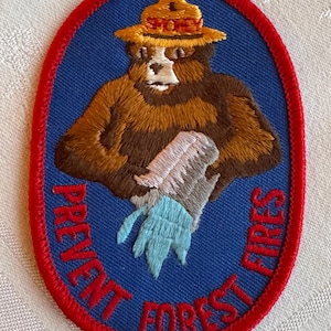 May include: An oval embroidered patch featuring a brown bear wearing a yellow ranger hat with the text SMOKEY. The bear holds a grey shovel with a light blue flame at the base. The blue background includes the red text PREVENT FOREST FIRES.