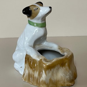 May include: A ceramic figurine of a dog, primarily white with brown and black markings, wearing a green collar. The dog is seated next to a brown tree stump-shaped container, possibly an ashtray or small planter.