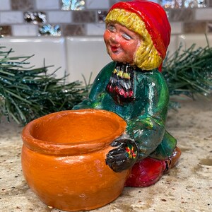 May include: A hand-painted ceramic figurine of a seated figure wearing a red hat, green coat, and red pants. The figure is holding a small, orange-brown pot. The figurine has a smiling face and is likely a decorative item.
