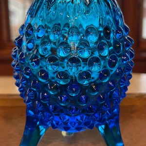 May include: A blue glass bud vase with a textured, bubbled surface and three legs. The vase is made of glass and has a teal blue color.