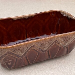 May include: A rectangular brown ceramic loaf pan with a textured interior and a decorative geometric pattern on the sides. The rim has a lighter, speckled finish. This baking dish is ideal for making bread or cakes.