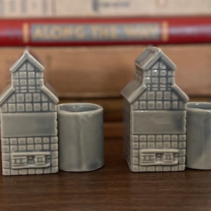 May include: Two gray ceramic house-shaped candle holders. Each has a rectangular body with a chimney and a cylindrical cup attached. The candle holders are on a wooden surface with books in the background.