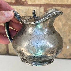 May include: A small, antique silver-colored creamer pitcher with a handle and a lid. The pitcher has a rounded body and a spout for pouring. The handle has a decorative design. The creamer is likely made of pewter or a similar metal.