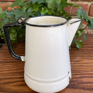 May include: White enamel pitcher with a black handle and spout. The pitcher has a black rim and a slight chip on the bottom.