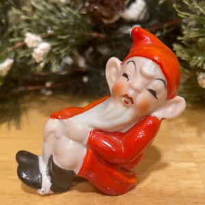 May include: A ceramic figurine of a sleeping gnome, wearing a red hat and coat with a white beard. The gnome has black shoes and is resting on its side. The background includes pine needles and a wooden surface.