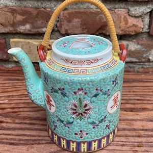 May include: A decorative ceramic teapot with a woven handle. The teapot features a turquoise background with floral and scroll patterns, red accents, and Chinese characters. The lid and base have colorful geometric designs.