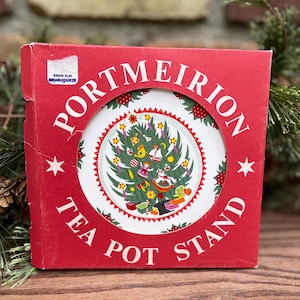 May include: A red box with the words "PORTMEIRION TEA POT STAND" in white. The tea pot stand is round with a Christmas tree design. The box has a Scandinavian Fifth Avenue sticker.