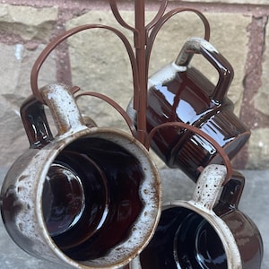 May include: Set of four brown and white ceramic mugs with a speckled glaze, displayed on a metal mug rack.