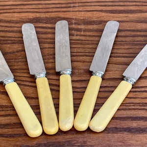 May include: Set of five butter knives with yellow plastic handles and silver blades.