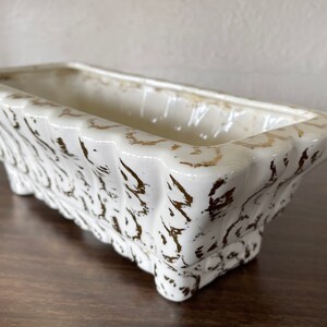 May include: Rectangular white ceramic planter with gold accents. The planter has a scalloped design on the sides and sits on small feet. The interior is a creamy white color, and the exterior has a distressed gold pattern.