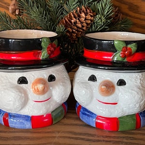 May include: Two ceramic snowman-shaped containers. Each has a white body, a carrot nose, black eyes, and a red smile. They wear a black hat with a red band and a scarf with blue, red, and green stripes. Holly and berries decorate the hat.
