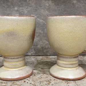 May include: Two ceramic mugs with handles, each with a footed base. The mugs are a light yellow color with brown and white marbling.