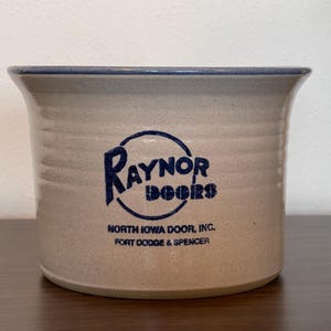 May include: A beige stoneware crock with a blue rim and a blue logo that reads "Raynor Doors". Below the logo is the text "North Iowa Door, Inc. Fort Dodge & Spencer". The crock has a cylindrical shape.