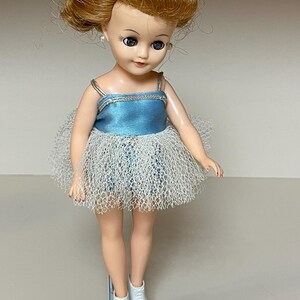 May include: A vintage doll with a blue satin bodice and a white tulle skirt. The doll has blonde hair and is wearing white ice skates. The doll is standing on a silver stand.
