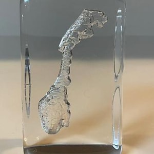 May include: A clear, rectangular glass paperweight with a detailed, three-dimensional depiction of the country of Norway. The map is rendered in a frosted, silvery finish, encased within the transparent glass. The paperweight sits on a light-colored surface.