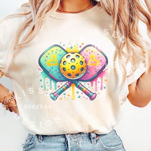 May include: A white t-shirt with a colorful graphic of two pickleball paddles crossed with a yellow pickleball in the center. The paddles have hearts on them and the graphic is surrounded by colorful paint splatters.