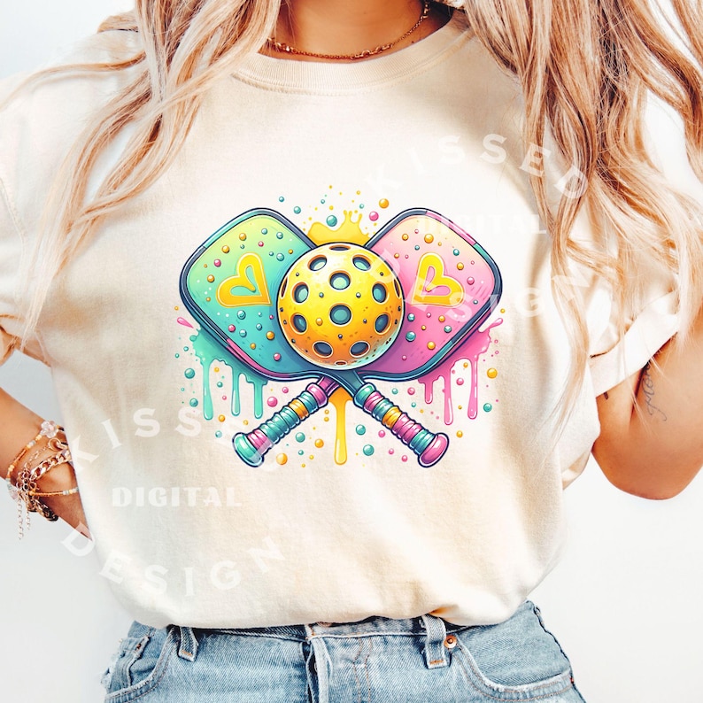 May include: A white t-shirt with a colorful graphic of two pickleball paddles crossed with a yellow pickleball in the center. The paddles have hearts on them and the graphic is surrounded by colorful paint splatters.