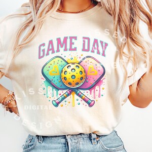 May include: A white t-shirt with the text "GAME DAY" in pink letters. The shirt features a graphic of two crossed pickleball paddles with a yellow pickleball in the center. The paddles are decorated with colorful drips and polka dots.