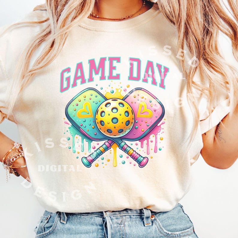 May include: A white t-shirt with the text "GAME DAY" in pink letters. The shirt features a graphic of two crossed pickleball paddles with a yellow pickleball in the center. The paddles are decorated with colorful drips and polka dots.