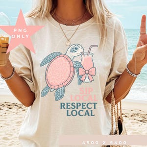 Peut inclure: T-shirt beige avec un motif de tortue marine rose et bleu, un cocktail et le texte "SIP LOCAL RESPECT LOCAL". La carapace de la tortue présente un motif mandala. Le t-shirt est de coupe décontractée.