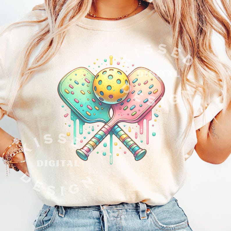 May include: A white t-shirt with a colorful graphic of two pickleball paddles crossed over a yellow pickleball. The paddles are decorated with colorful sprinkles and dripping with colorful icing.