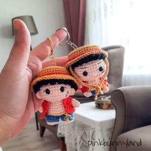 May include: Two crochet keychains featuring a cartoon character with a straw hat, red vest, and blue shorts. The keychains are held by a hand. The keychains have black eyes and a smiling face. The keychains are attached to a gold-colored keychain ring.