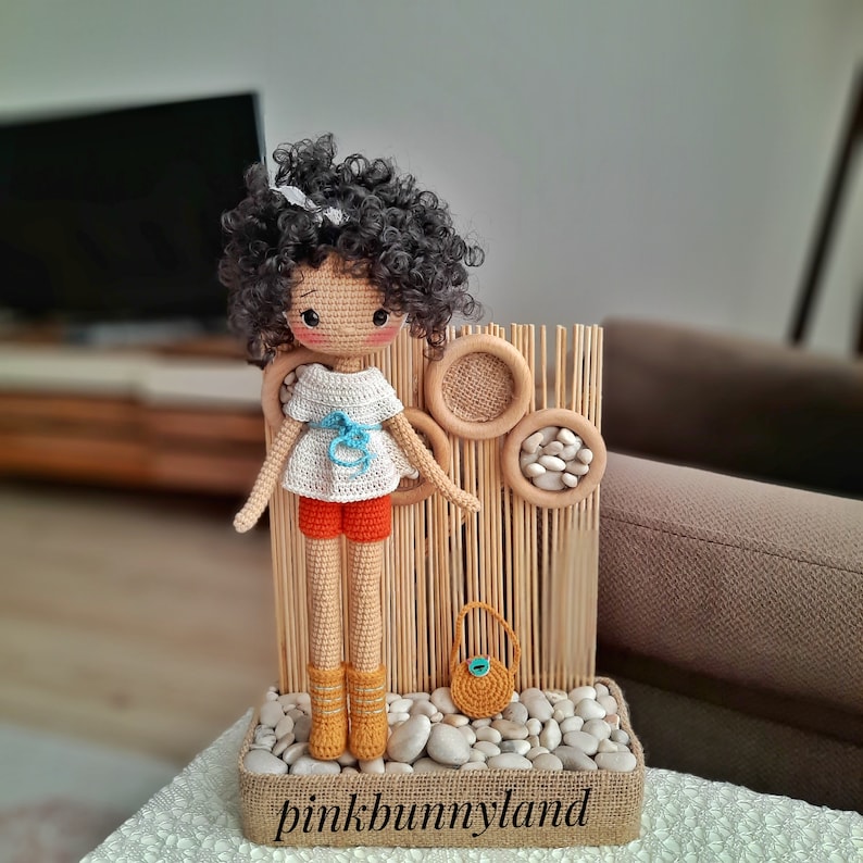 Op de afbeelding: Een gehaakte pop met krullend bruin haar, die een wit shirt met een blauwe strik, een oranje broekje en gele laarzen draagt. De pop staat voor een houten achtergrond met een klein bruin tasje.