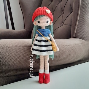May include: A crocheted doll wearing a red and white striped dress, a blue top, and a green and yellow purse. The doll has a red hat with a white heart on it and green hair.