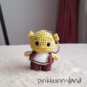 Puede incluir: Un llavero de ganchillo hecho a mano con un personaje de ogro verde con cabeza amarilla, chaleco y pantalones marrones y camisa blanca. El llavero tiene un anillo plateado. El texto "pinkbunnyland" es visible.