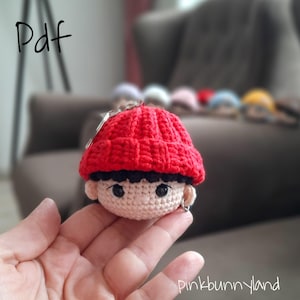 May include: A crocheted doll head with a red beanie, black hair, and small silver hoop earrings. The doll's face is beige with black eyes. The word "Pdf" is in the upper left corner. The text "pinkbunnyland" is in the lower right corner.