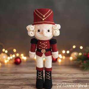May include: A crocheted nutcracker doll featuring a deep red hat with gold trim, a matching red jacket with gold cross-stitching, white trousers, and black boots with gold laces. The figure has curly white hair and a white mustache.
