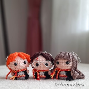 May include: Three handmade crochet character keychains. The keychains feature characters with orange, brown, and brown hair, each wearing a scarf with red and gold stripes. The characters are wearing black robes and have black eyes. The keychains are on a white surface.