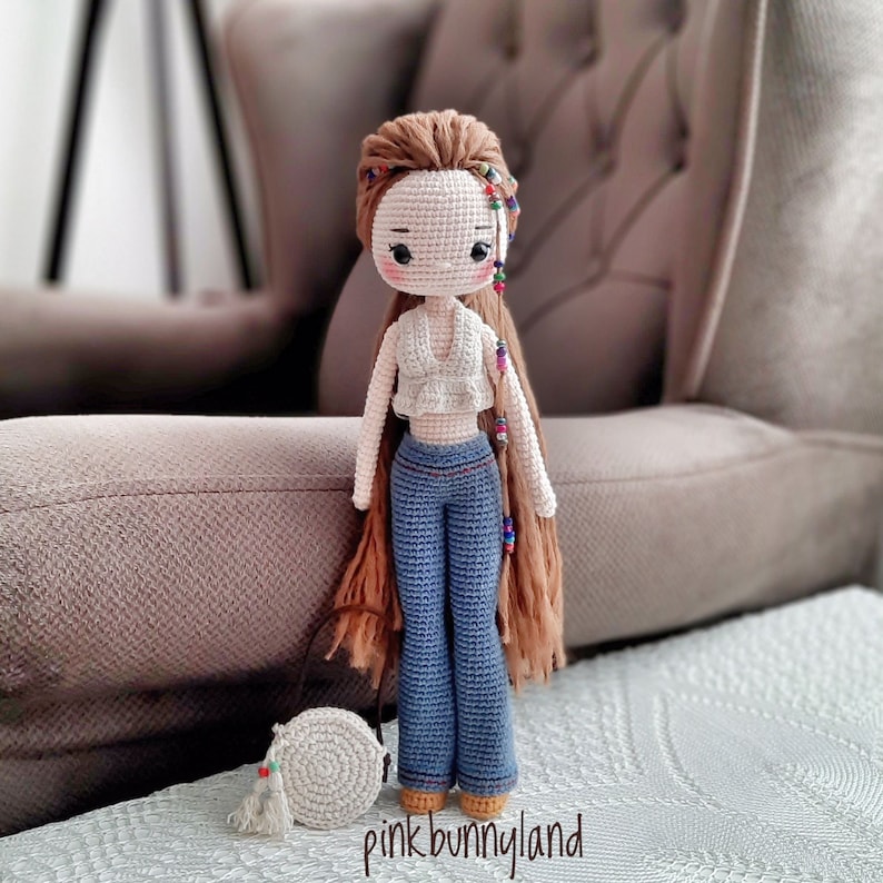 Puede incluir: Una mu&ntilde;eca de crochet con el pelo largo y marr&oacute;n, que lleva un top blanco corto y unos vaqueros azules. La mu&ntilde;eca lleva una peque&ntilde;a bolsa marr&oacute;n con un c&iacute;rculo blanco.