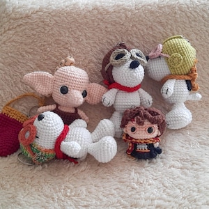 May include: A collection of handmade crochet character keychains. The keychains include a pink-eared creature, a white dog with aviator goggles and a red scarf, a white dog with a green hat, and a small Harry Potter figure.